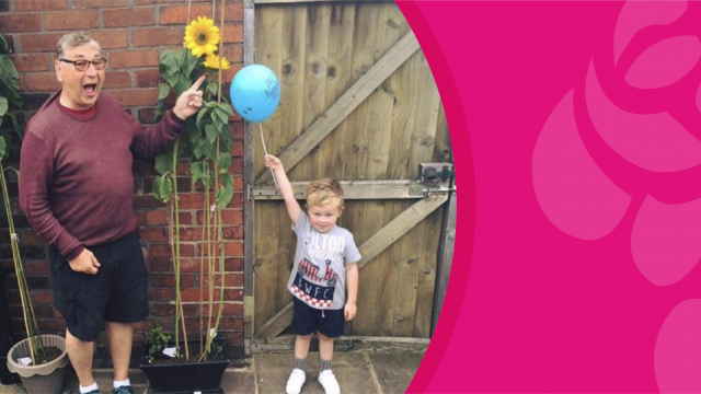 Young boy and man stood either side of a tall sunflower in a back garden.