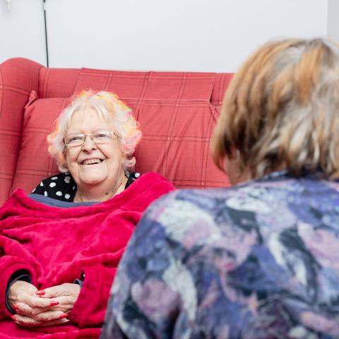 Elderly woman sat in chair, talking to a member of hospice staff.